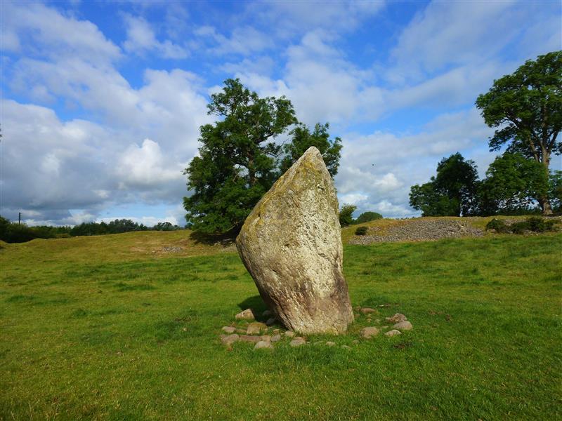 A close up photo of the standing stone at Mayburgh Henge, Cumbria, England. The stone rises to a point and has irregular sides. It is surrounded by smaller stones at its base. In the background you can see part of the henge mound that encloses the stone, along with several trees. A close up photo of the standing stone at Mayburgh Henge, Cumbria, England. The stone rises to a point and has irregular sides. It is surrounded by smaller stones at its base. In the background you can see part of the henge mound that encloses the stone, along with several trees.