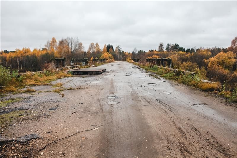 A photo of an abandoned bridge leading into the ghost town of Centralia where coal fires have been burning since 1962. The road is damp from rain and has pot holes, plant growth and debris covering it. The sides of the bridge have autumn trees of various shades of yellow and brown and the sky is a thick grey.
