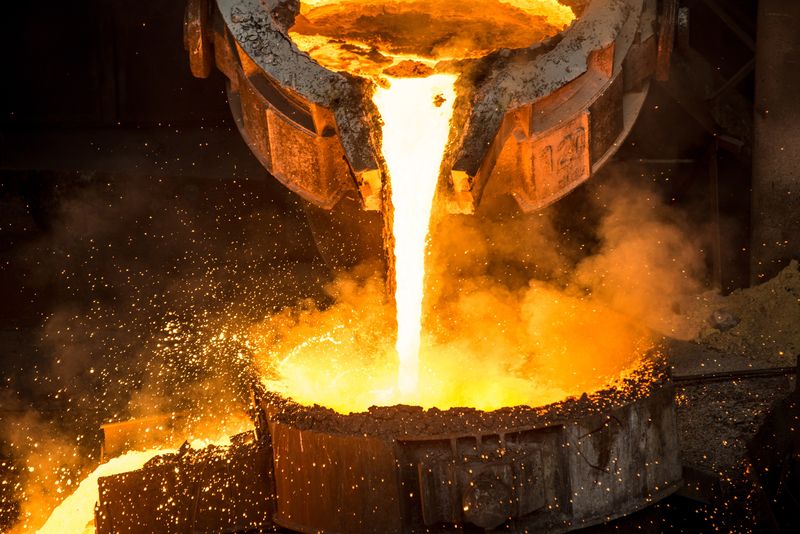 A ladle pouring molten metal that looks orange like it's on fire into another poth, sparks are flying up all around. 