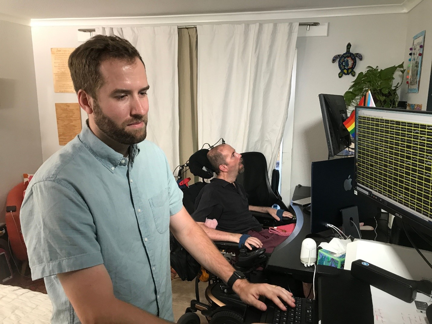 Postdoctoral scholar and lead author of the study Nicholas Card getting the BCI system ready. Card is in the foreground, a white man with brown hair and bear wearing a pale blue short sleeved shirt. He is standing in front of a computer. In the background Casey Harrell, a white man who uses an electric wheelchair, is positioned in front of another computer monitor. Postdoctoral scholar and lead author of the study Nicholas Card getting the BCI system ready. Card is in the foreground, a white man with brown hair and bear wearing a pale blue short sleeved shirt. He is standing in front of a computer. In the background Casey Harrell, a white man who uses an electric wheelchair, is positioned in front of another computer monitor.
