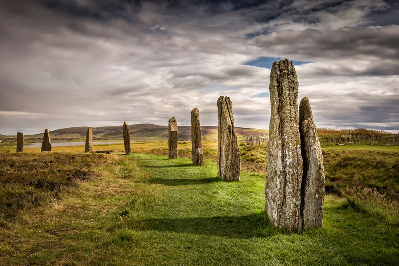 A close up photo of the standing stones at the Ring of Brodgar site in Orkney. The photo shows eight stones going into the distance as they mark out the circle of the structure. The stones sit on a band of grass which is surrounded by more unkept grasses. The sky is overcast with small slivers of blue.  