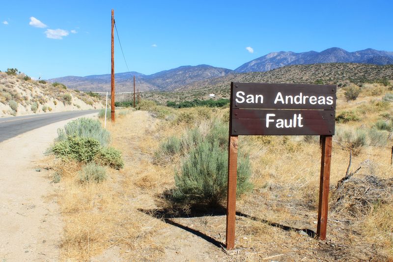A sign saying "San Andreas Fault", standing in a dry, grassy area with hills in the background.