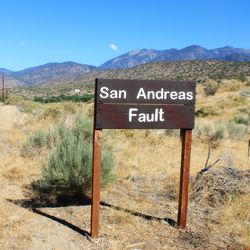 A sign saying "San Andreas Fault", standing in a dry, grassy area with hills in the background.