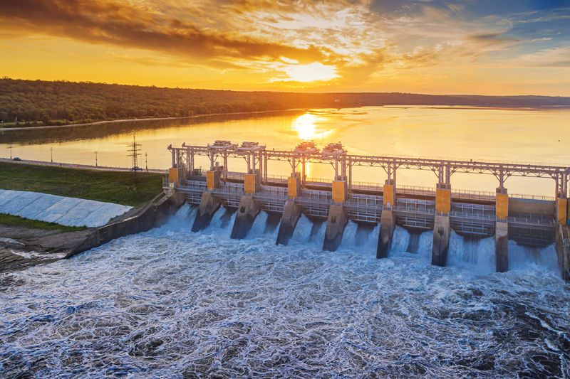 A photo of a hydroelectric power station at sunset. The Water is cascading through a number of channels. The water behind the dam is golden as it reflects the sun. 