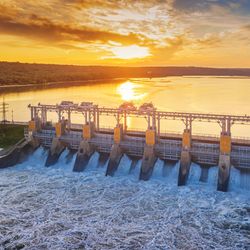 A photo of a hydroelectric power station at sunset. The Water is cascading through a number of channels. The water behind the dam is golden as it reflects the sun. 