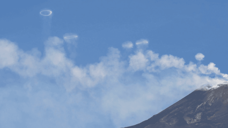On the right hand side is the top of Mount Etna and the rest of the image is blue sky and white vapor. Above the white vapor are perfect smoke-like rings.